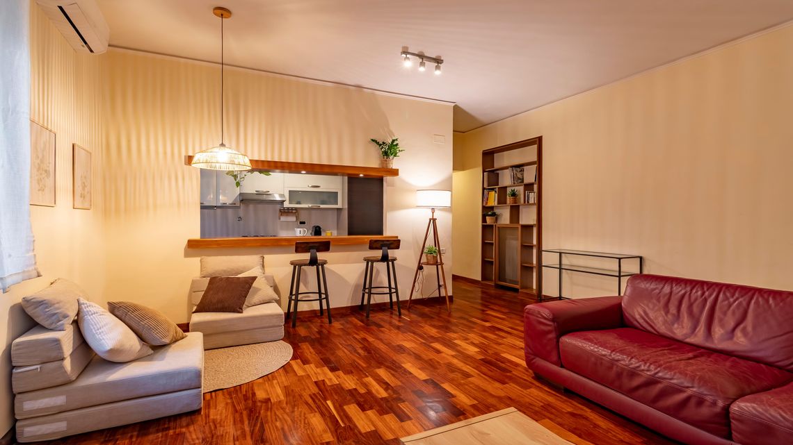 Il soggiorno dell'appartamento a Cornelia: parquet rosso, divano bordeaux, poltrone chiare e la cucina a vista