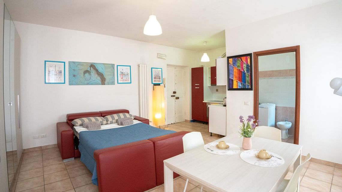 Il monolocale visto dalla porta: il letto, il tavolo apparecchiato con i fiori, i quadri alle pareti