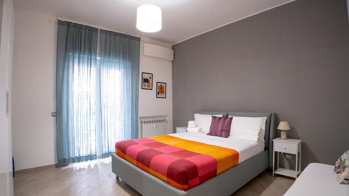 La camera dell'appartamento al Pigneto: parete grigia, letto con il copriletto a quadri rossi e arancioni, tende azzurre