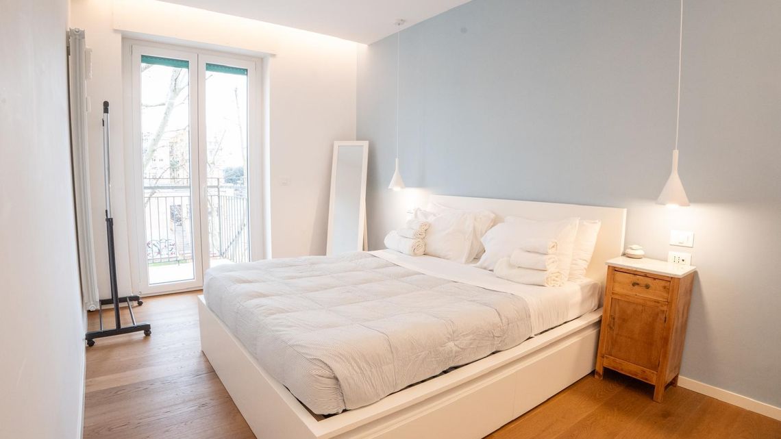 La camera: letto bianco, comodino di legno e la portafinestra sul balcone